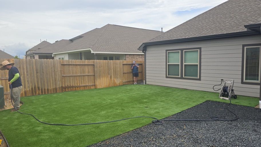 Two workers installing artificial turf in a backyard with a wooden fence and house.