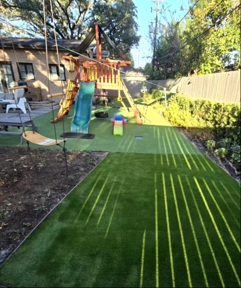 Backyard with playground on artificial turf, sunlit shadows. Wooden play structure, slide, swing set.