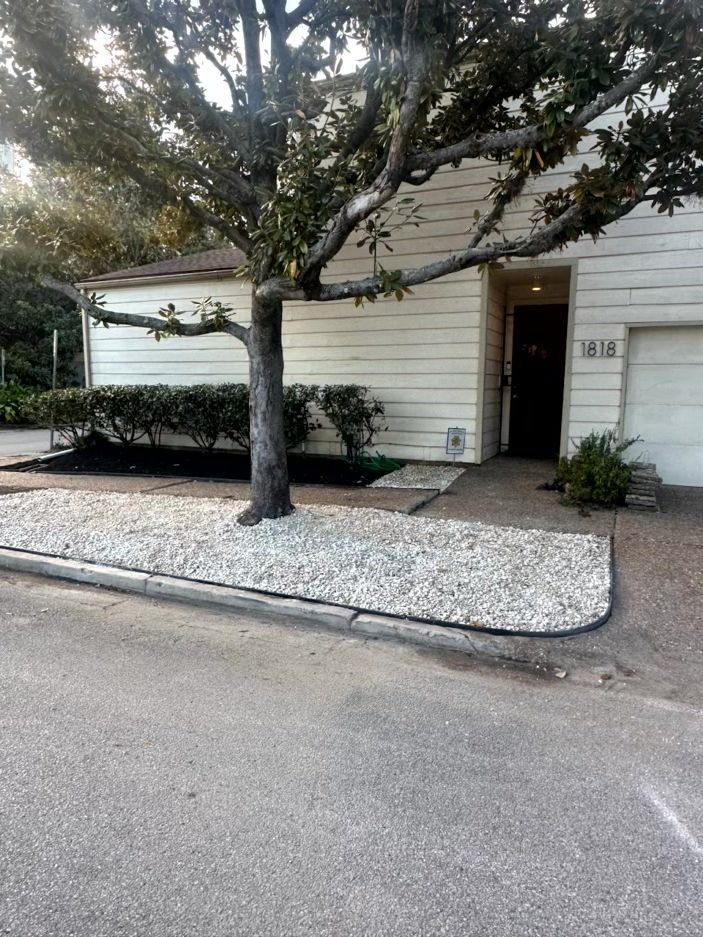 A beige house with a tree in front. White gravel, bushes, and a pathway lead to the front door.