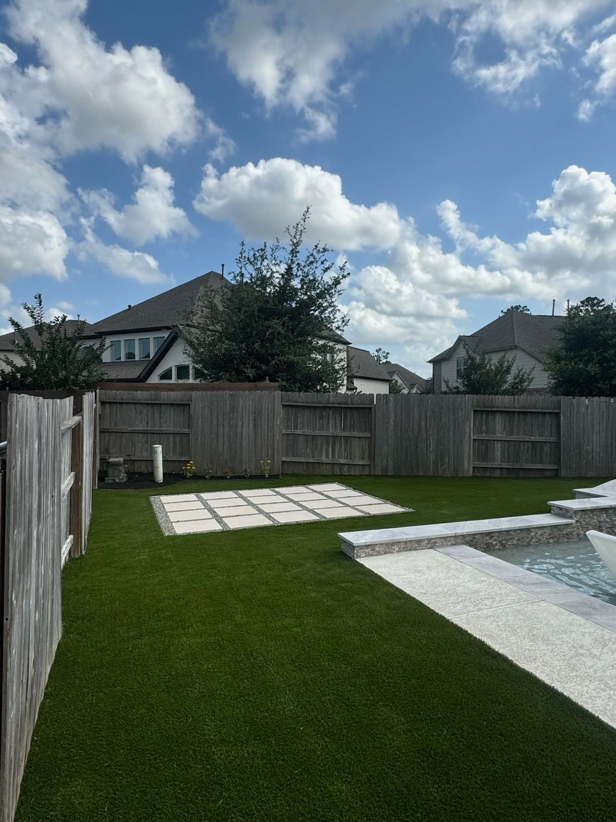Backyard with green lawn, wooden fence, pool, and houses under a cloudy sky.