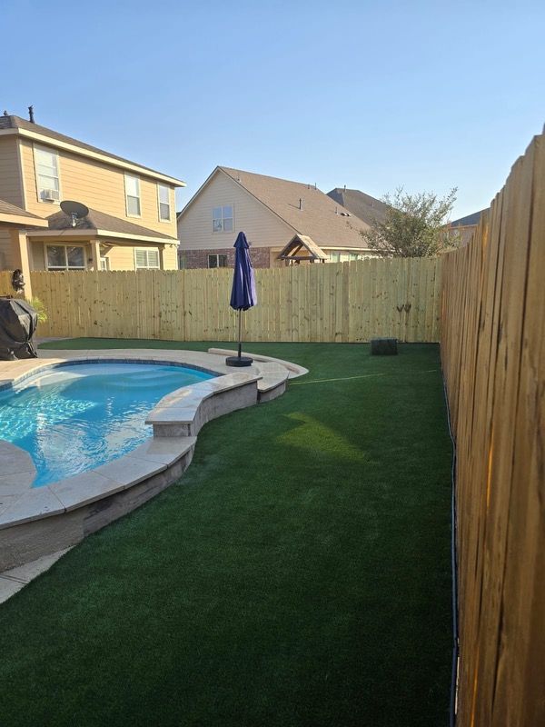 Backyard with a swimming pool, artificial turf, and a wooden fence; blue umbrella stands.