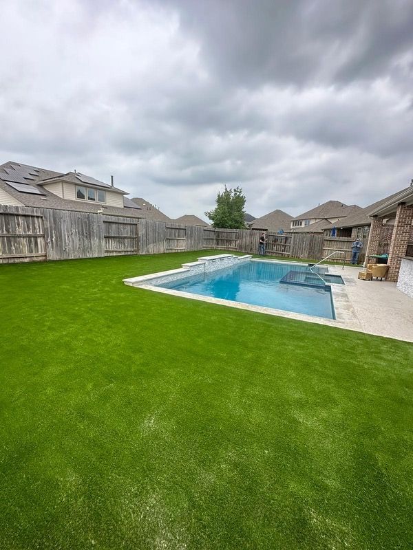 Backyard with rectangular pool, surrounded by green turf and a wooden fence under a cloudy sky.