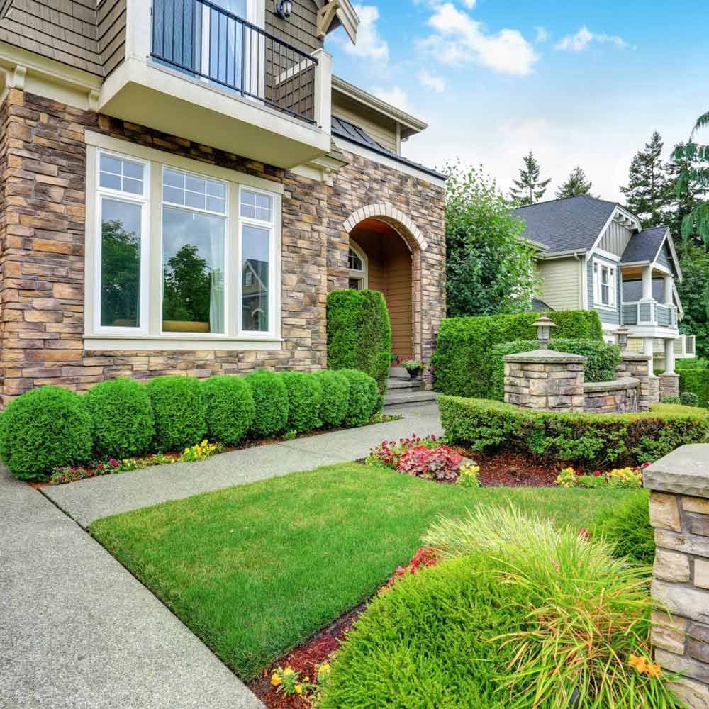 Luxurious stone-clad home with manicured lawn, shrubs, and neighboring houses under a blue sky.