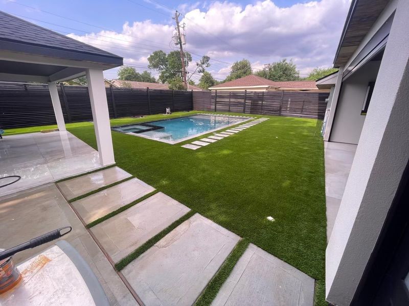 Backyard with pool, patio, and green lawn. Light stone walkways. Black fence.