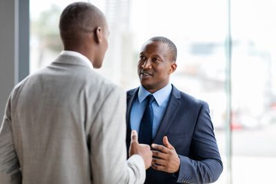 Two businesspeople in suits talking in a bright office, one gesturing while speaking