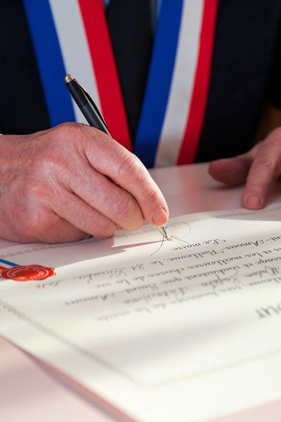 Hand signing a document with a pen beside a red seal and blue-red-white tie