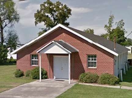 Small red brick house with a white front door, gabled porch, and concrete driveway.