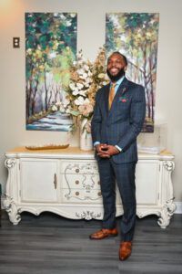 Man in a navy suit standing in a room beside a white cabinet and floral arrangement