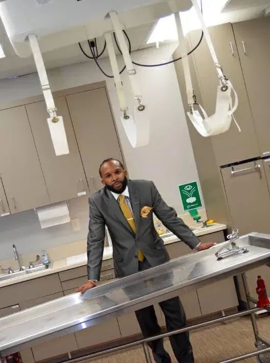 A man in a gray suit stands in a medical exam room beside a stainless steel table.