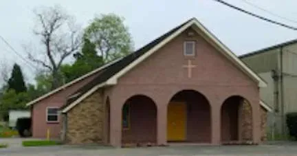 Small pink church with arched porch openings, a cross on the front, and a stone side wall