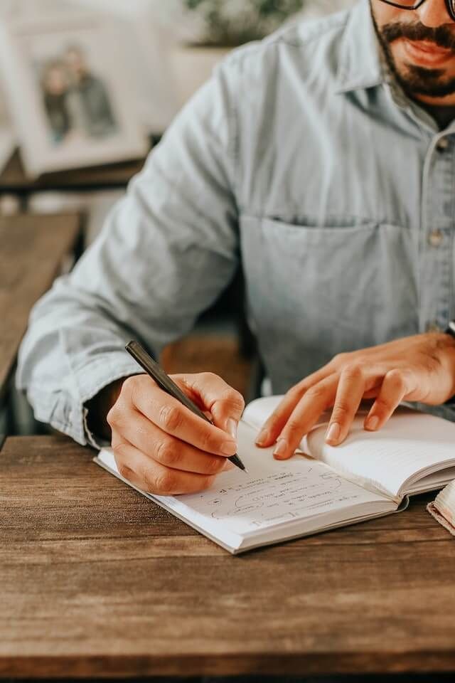 Person writing in a notebook at a wooden table, with another hand pointing at the page.