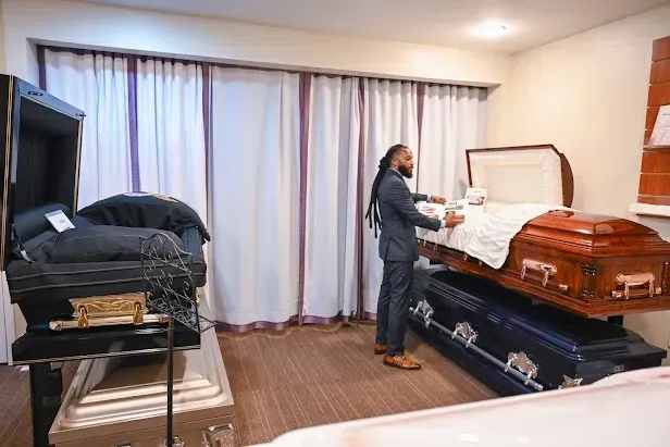 Person arranging a brown casket in a room with another casket nearby and closed curtains in the background