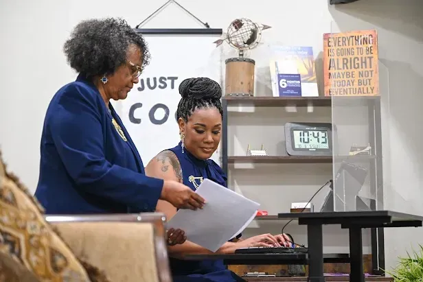 Two women reviewing papers at a desk in an office, with a laptop and shelves in the background.