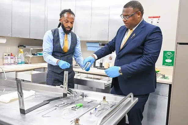 Two men in lab coats and gloves examine equipment on a stainless steel worktable in a lab.
