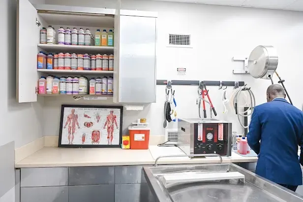 Clinic exam room with medical supplies on shelves and a person in blue working at a sink and equipment