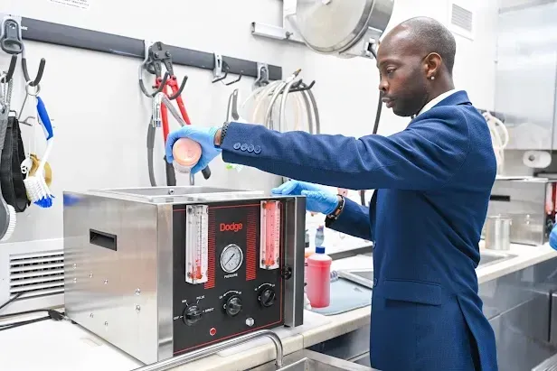 Person in blue coat using a lab machine with a gloved hand in a stainless-steel workshop