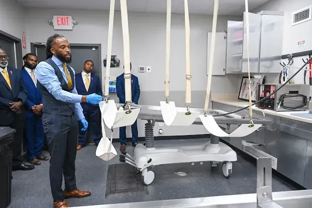 Men inspecting a white hospital bed frame in an industrial workshop with machinery nearby