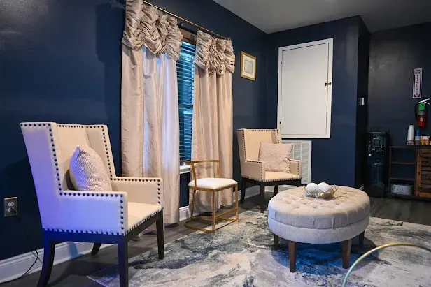 Blue living room with two beige armchairs, round ottoman, patterned rug, and curtained window