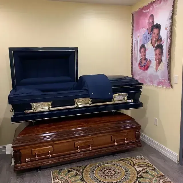 Two stacked caskets in a beige room, one black open casket above a brown one, with a memorial photo on the wall.