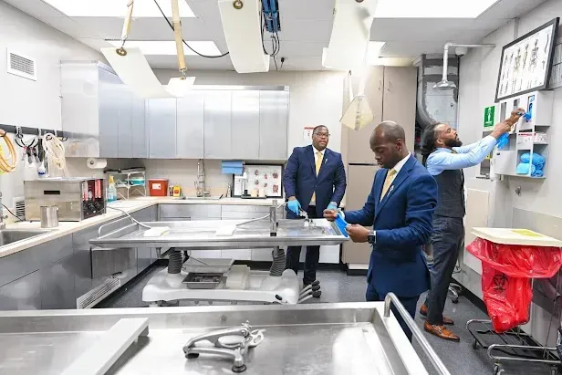 People in blue blazers in a stainless-steel lab, one pointing at wall controls near a sink area.