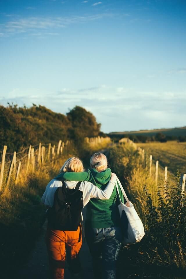 Two hikers with backpacks walking down a grassy path beside tall reeds under a blue sky