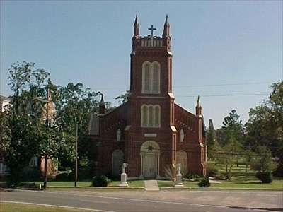 Red-brick church with twin towers and a cross, centered in a grassy yard under a clear sky.