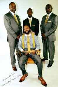 Four men in coordinated suits pose in a studio, one seated in front with yellow tie and brown shoes.