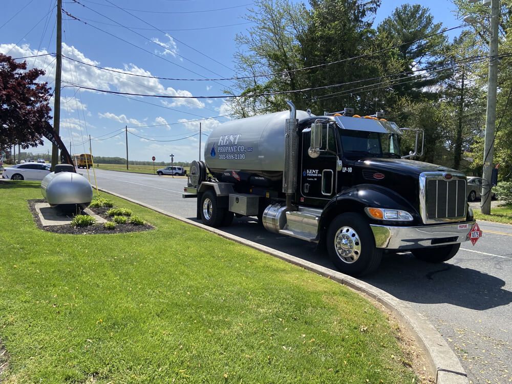 Black tanker truck parked by a curb on a sunny roadside with grass and trees.