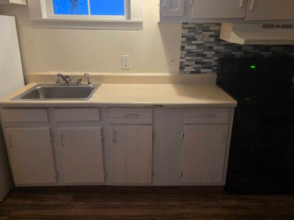 Small kitchen with white cabinets, sink under a window, tiled backsplash, and a black appliance.