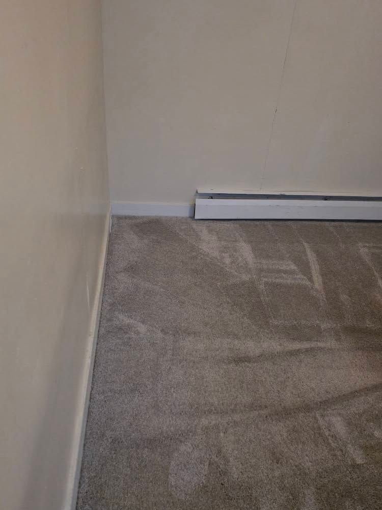Empty carpeted room corner with beige walls and white baseboard heating unit.