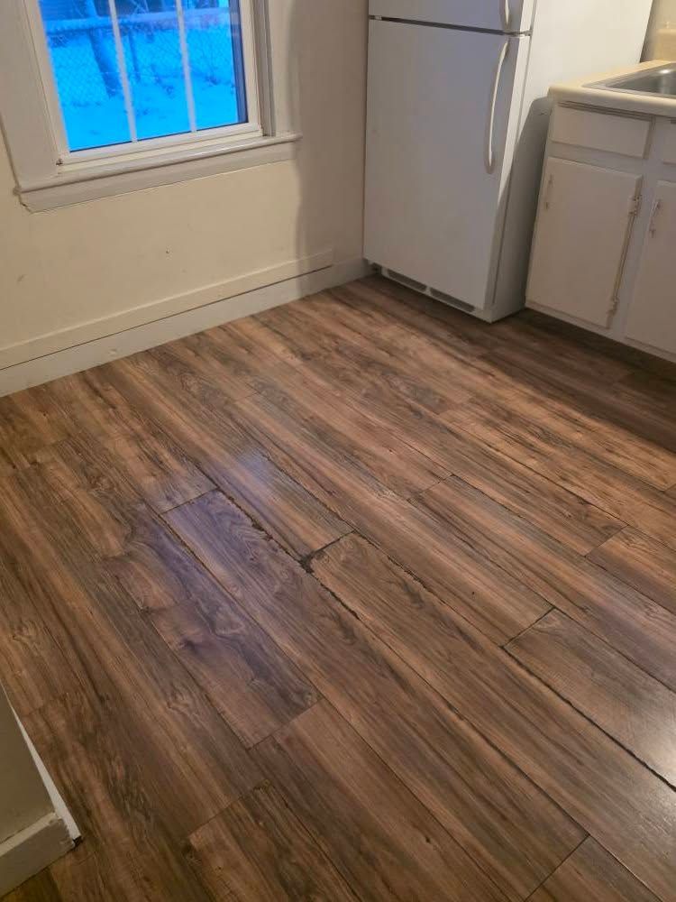 Empty kitchen corner with wood-look flooring, white cabinets, a refrigerator, and a window.
