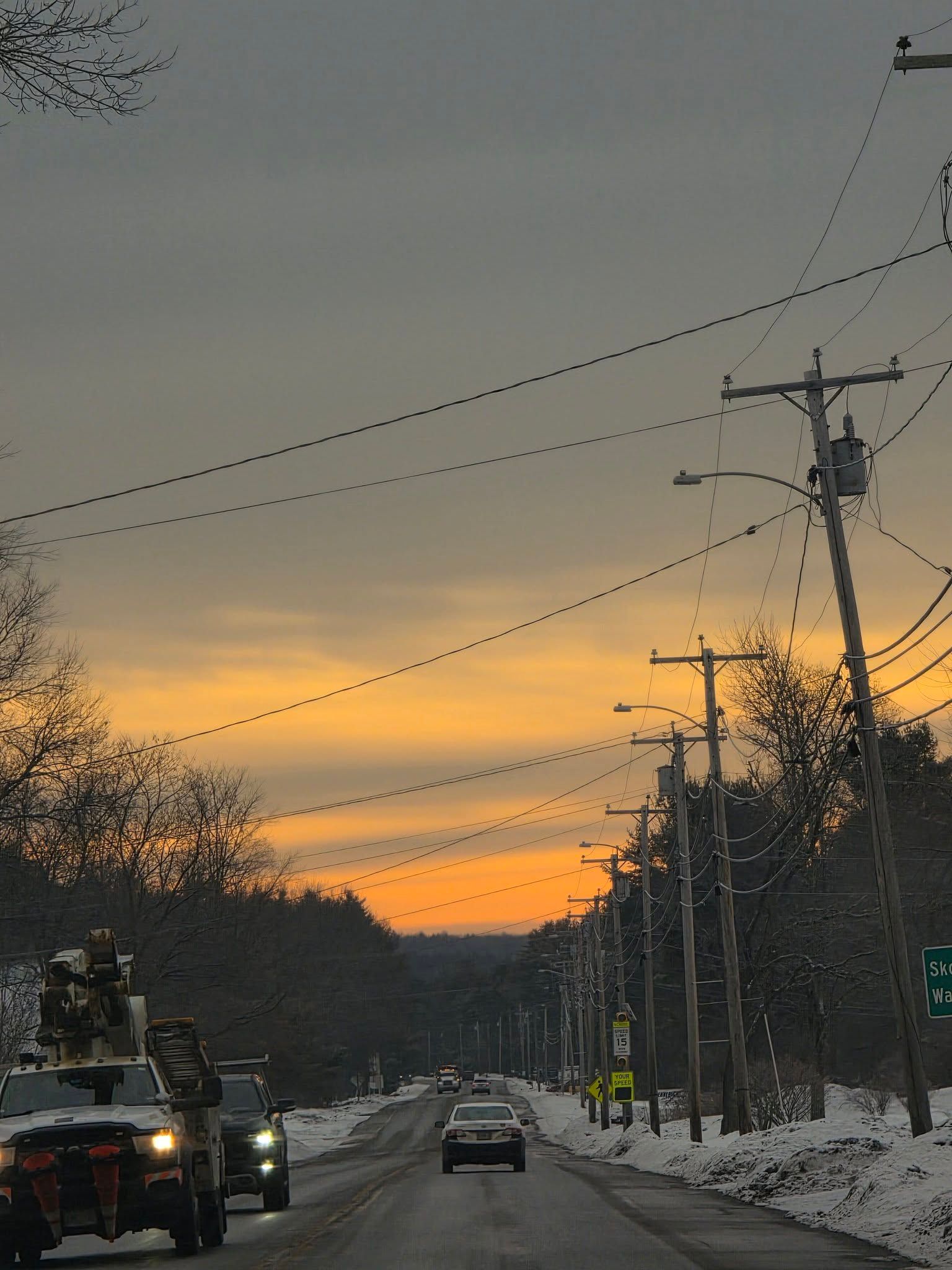 Snowy road at dusk with cars, utility poles, and a warm orange sunset sky