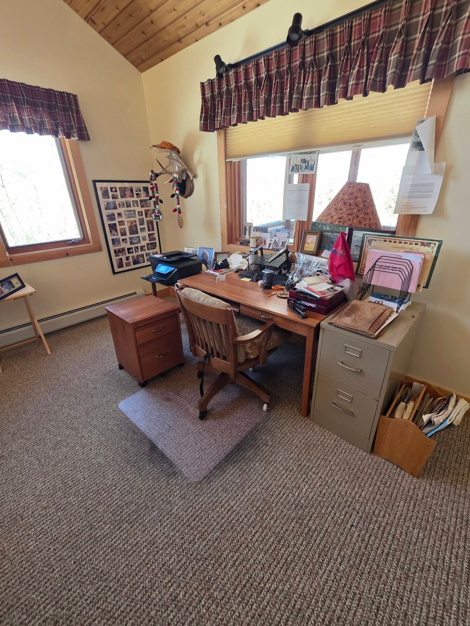 Home office with a wooden desk, computer, lamp, chair, and filing cabinets in a carpeted room with high windows.