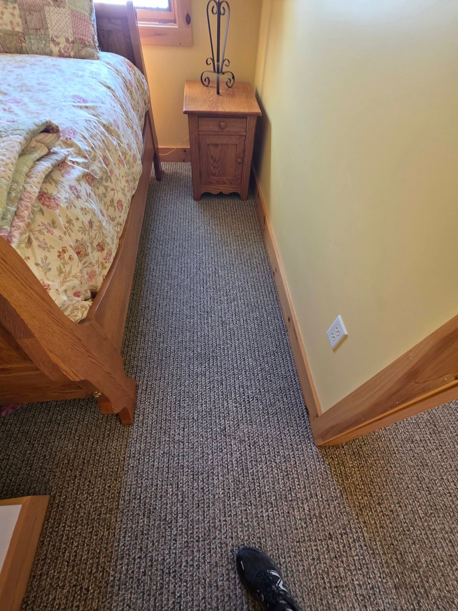 Bedroom corner with bed, nightstand, beige walls, carpet, and a shoe visible on the floor.