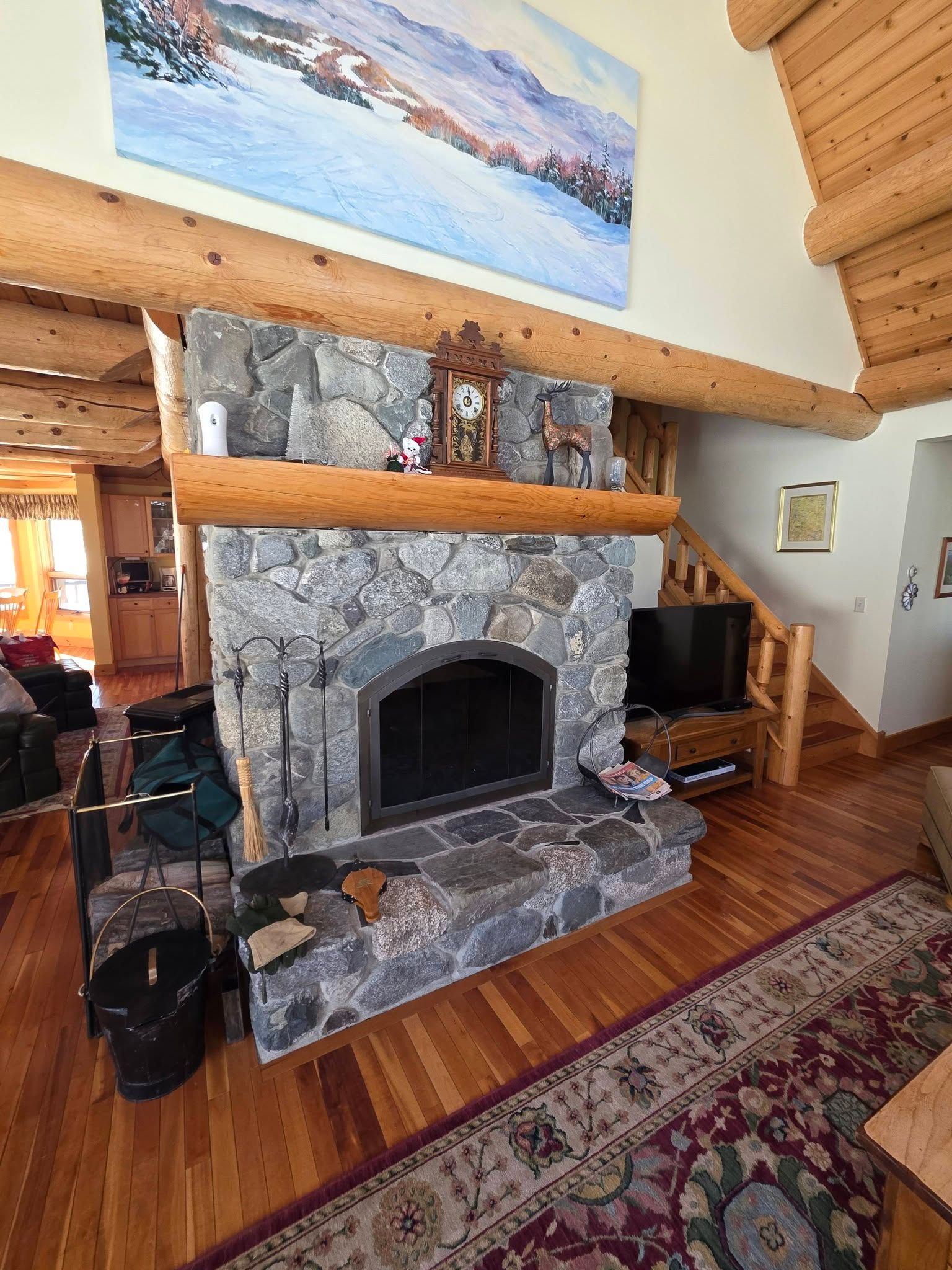 Rustic stone fireplace in a log cabin living room with wood floors and a landscape painting above.