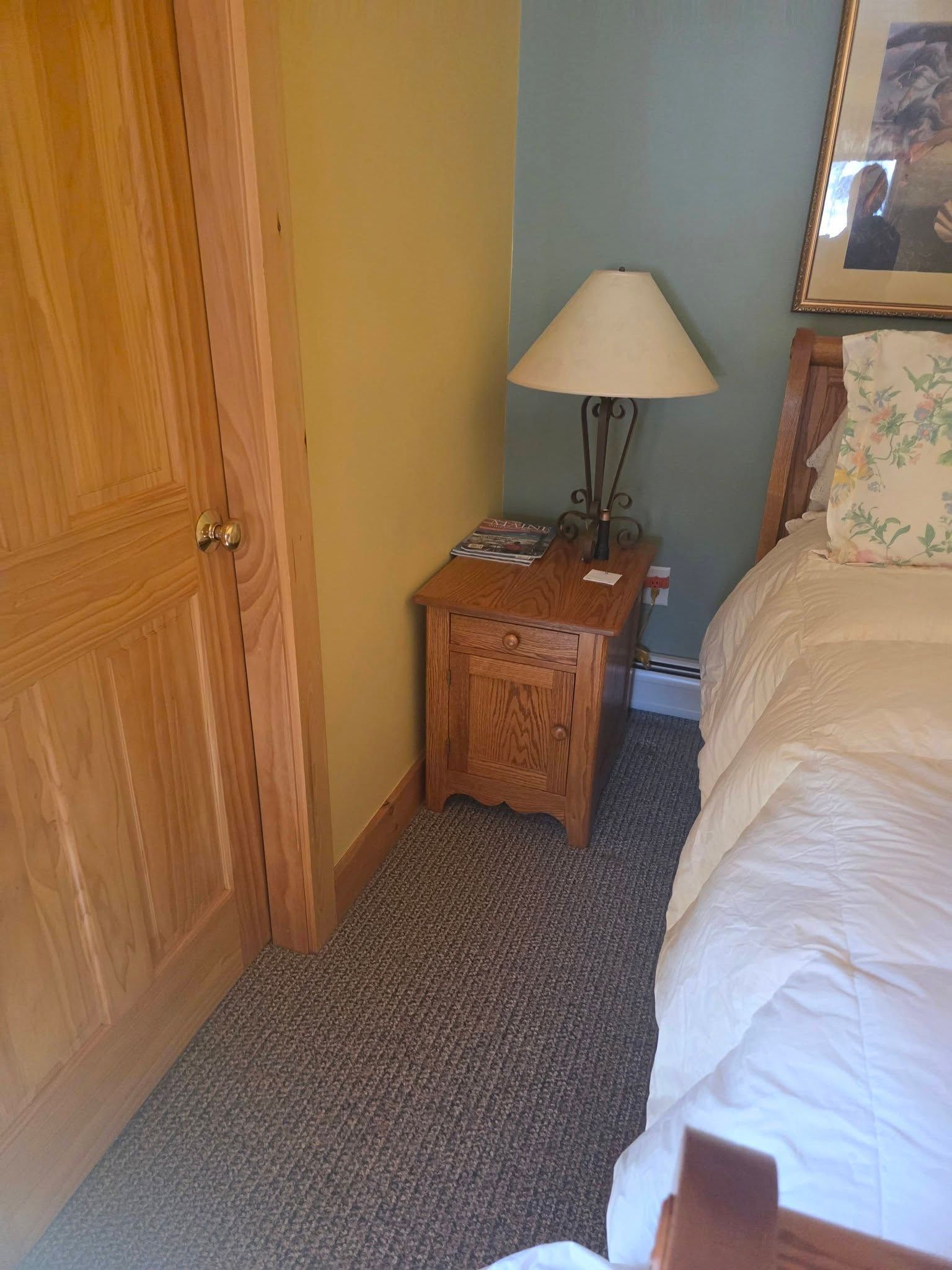 Bedroom corner with wooden nightstand, lamp, closet doors, and bed with white bedding.