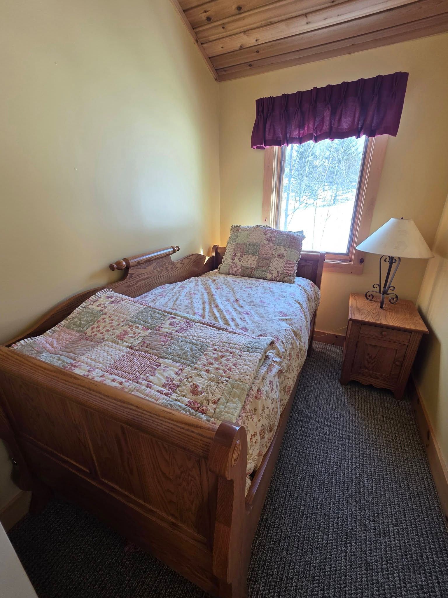 Small bedroom with wooden bed, floral bedding, purple curtains, and a bedside lamp.