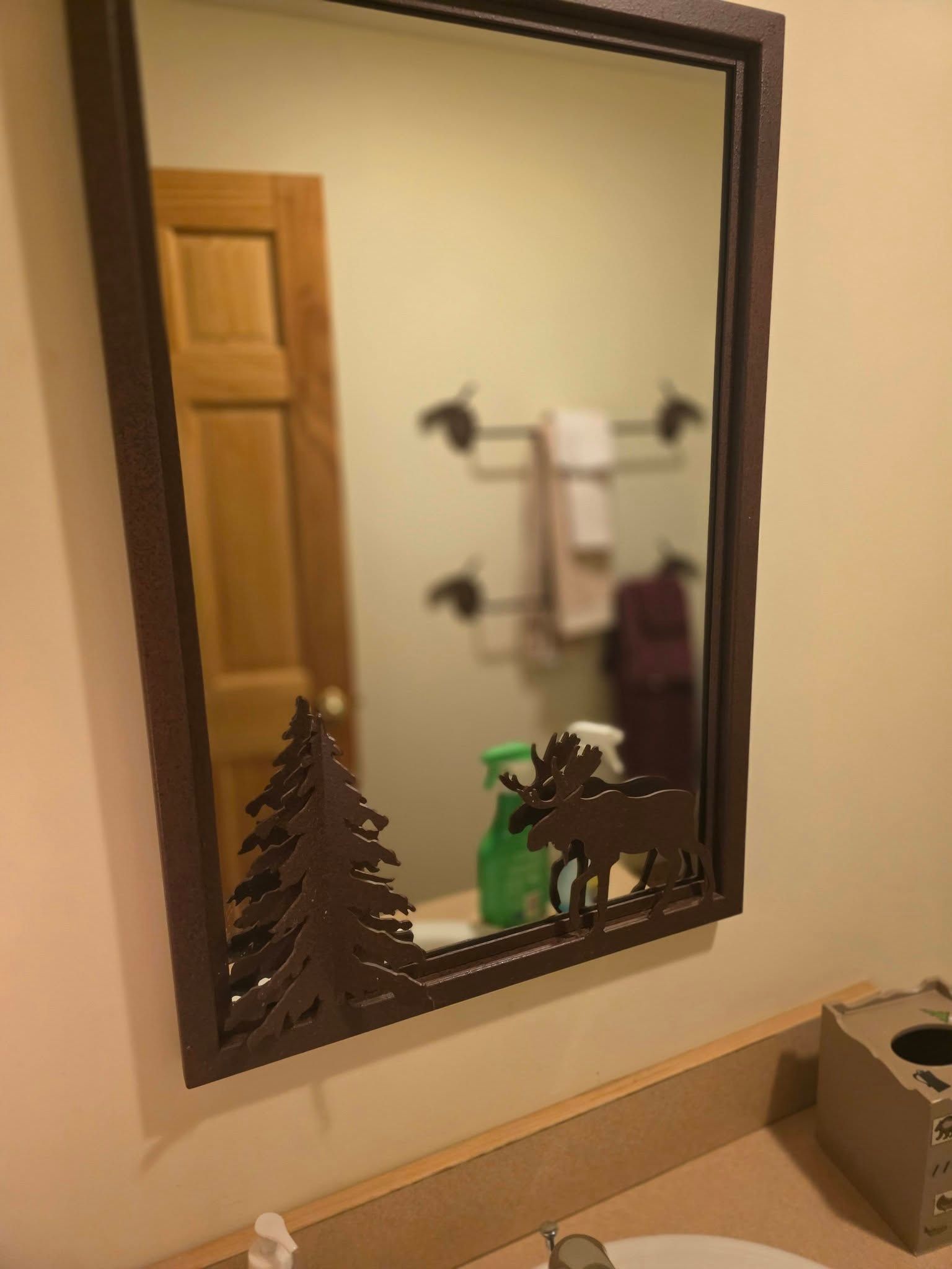 Bathroom mirror reflecting a towel rack, wooden door, and small pine tree and moose decorations on the counter.