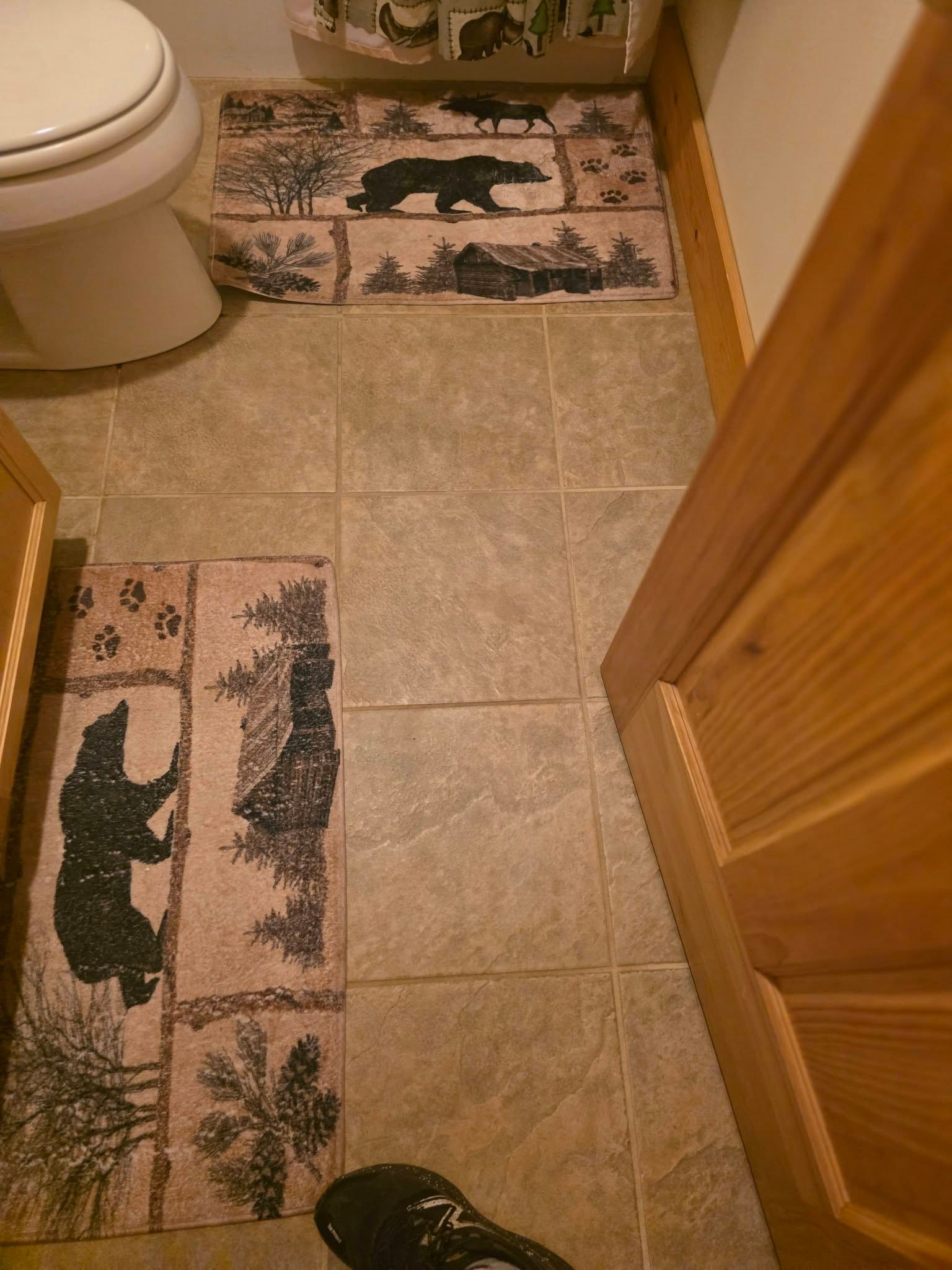 Small bathroom with beige tile floor, toilet, open wooden door, and two patterned rugs.