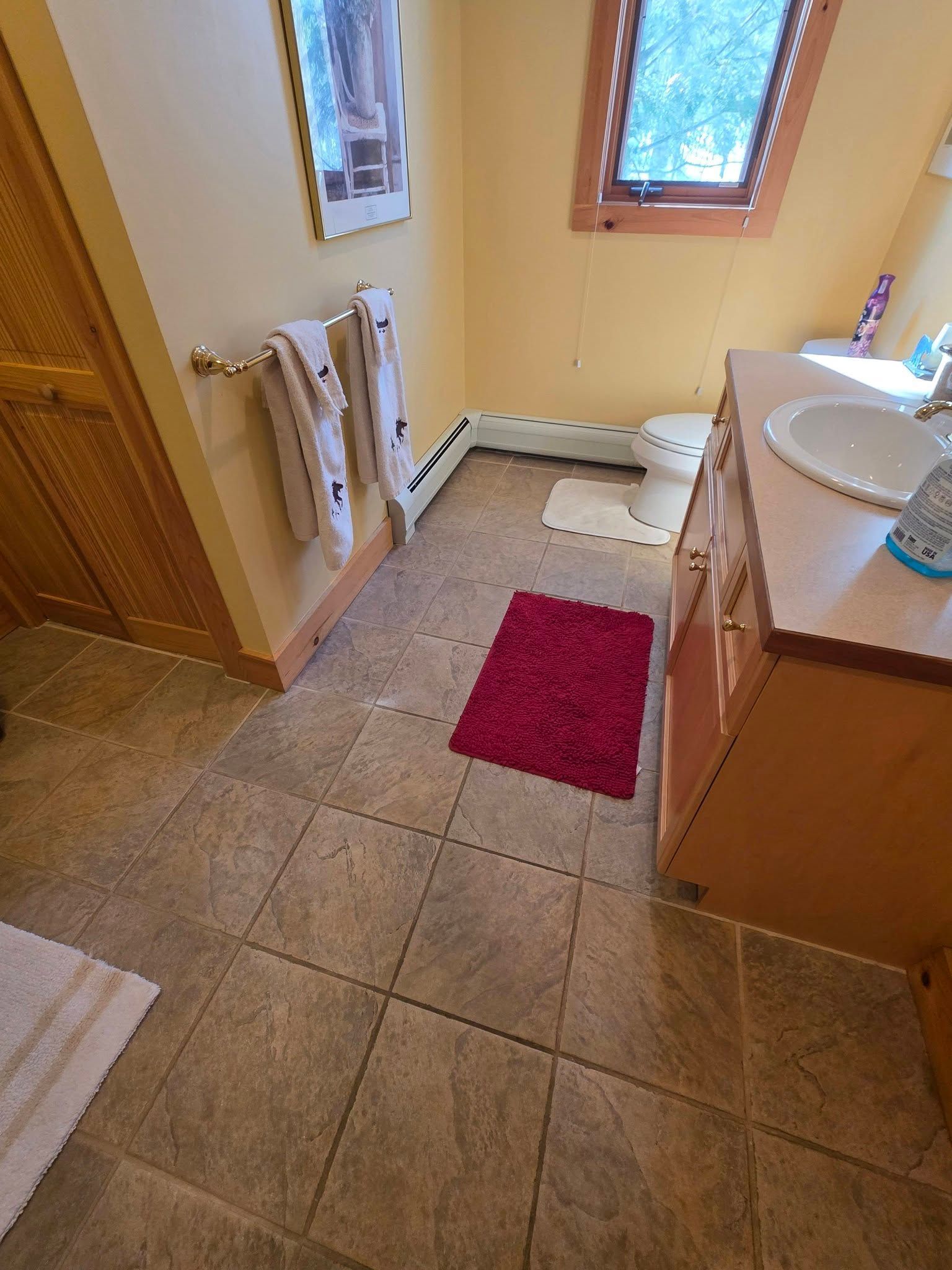 Small bathroom with beige tile, white sink and toilet, window, radiator, and red bath mat.