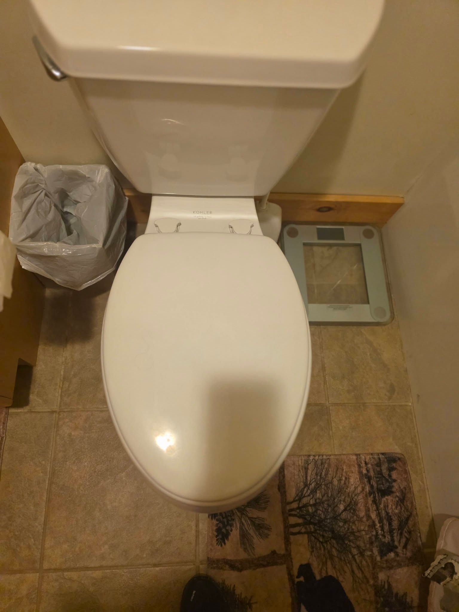 Top-down view of a toilet in a small beige-tiled bathroom with a trash can and floor scale nearby