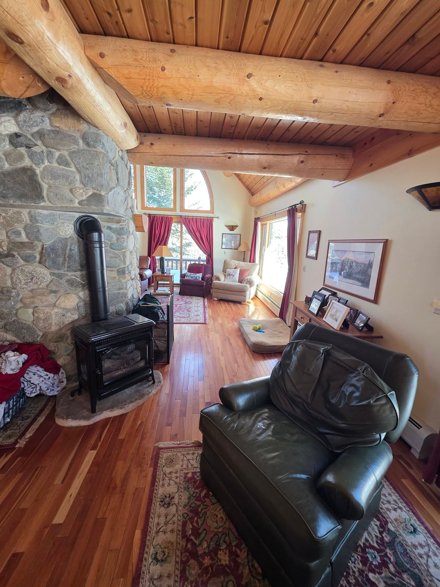 Cozy log-cabin living room with stone fireplace, leather chair, and wood-beamed ceiling overlooking a bright doorway.