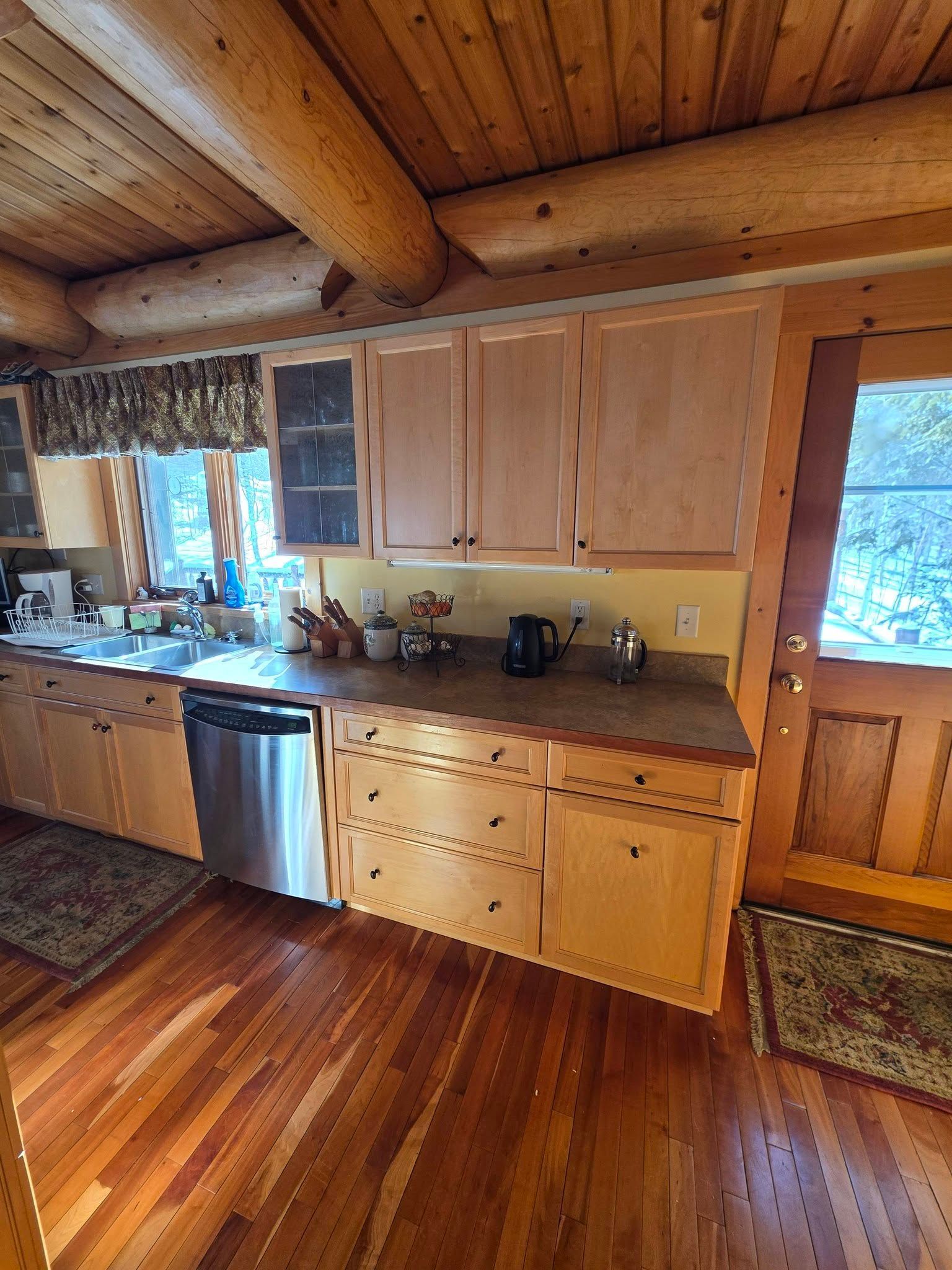 Rustic kitchen with wood-paneled walls, cabinets, stainless dishwasher, and a window over the sink
