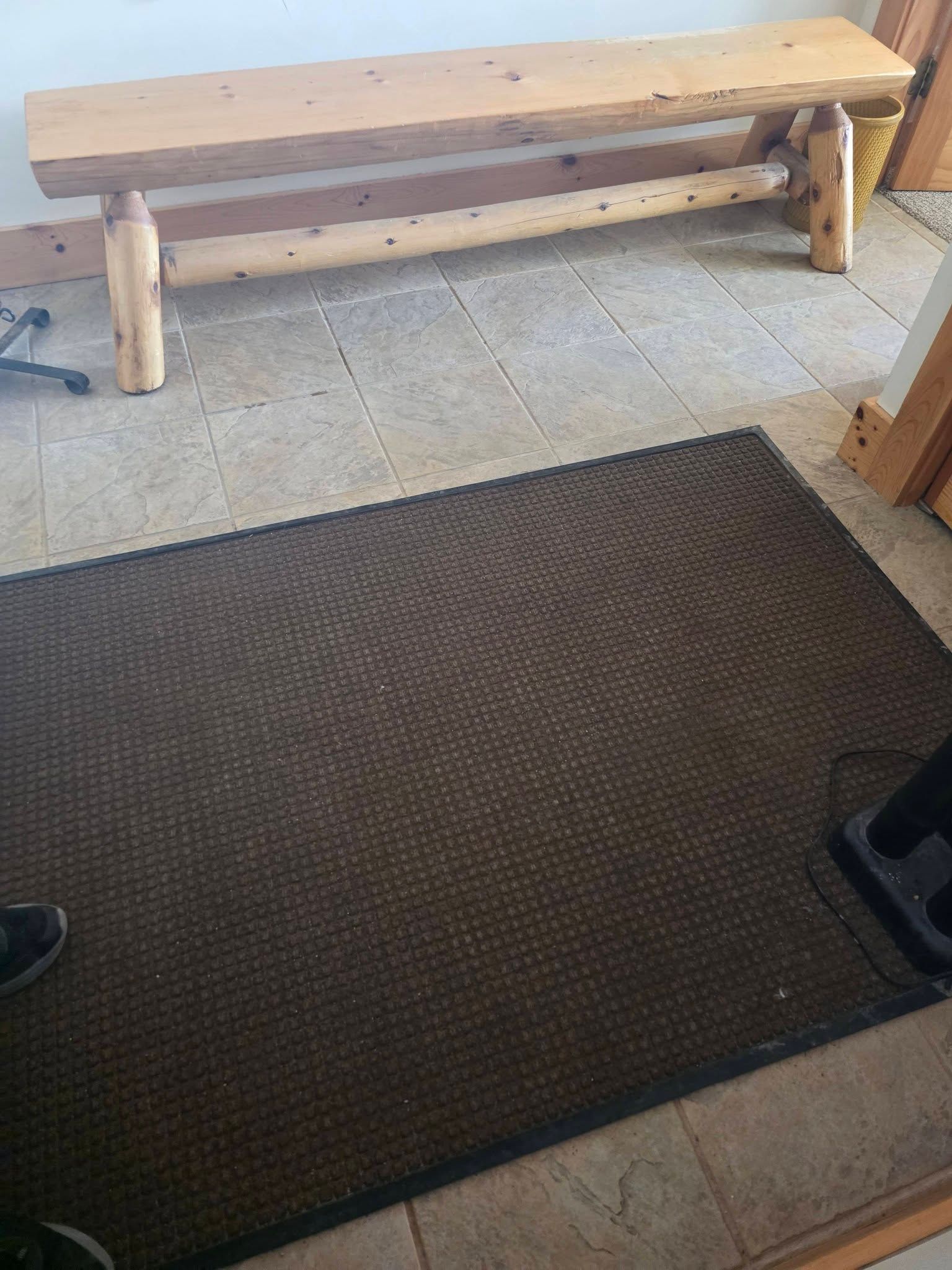 Wooden bench on a tiled floor beside a dark brown mat in an indoor entryway.