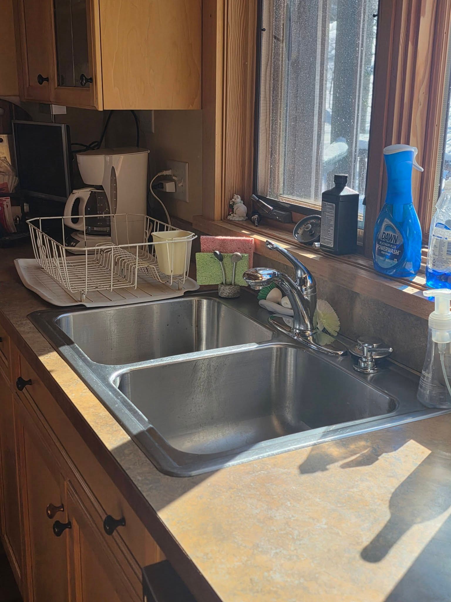 Kitchen sink beside a sunlit window with dishes, soap, and a blue bottle on the counter