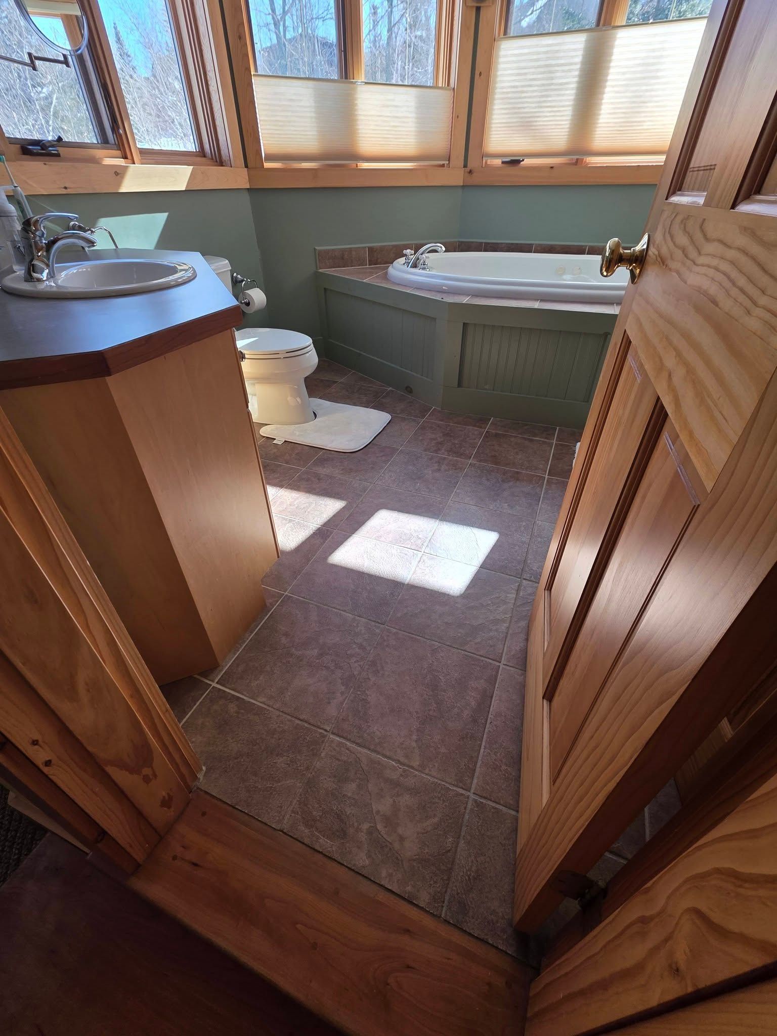 Small bathroom with a green corner bathtub, toilet, sink, and sunlight on tile floor.