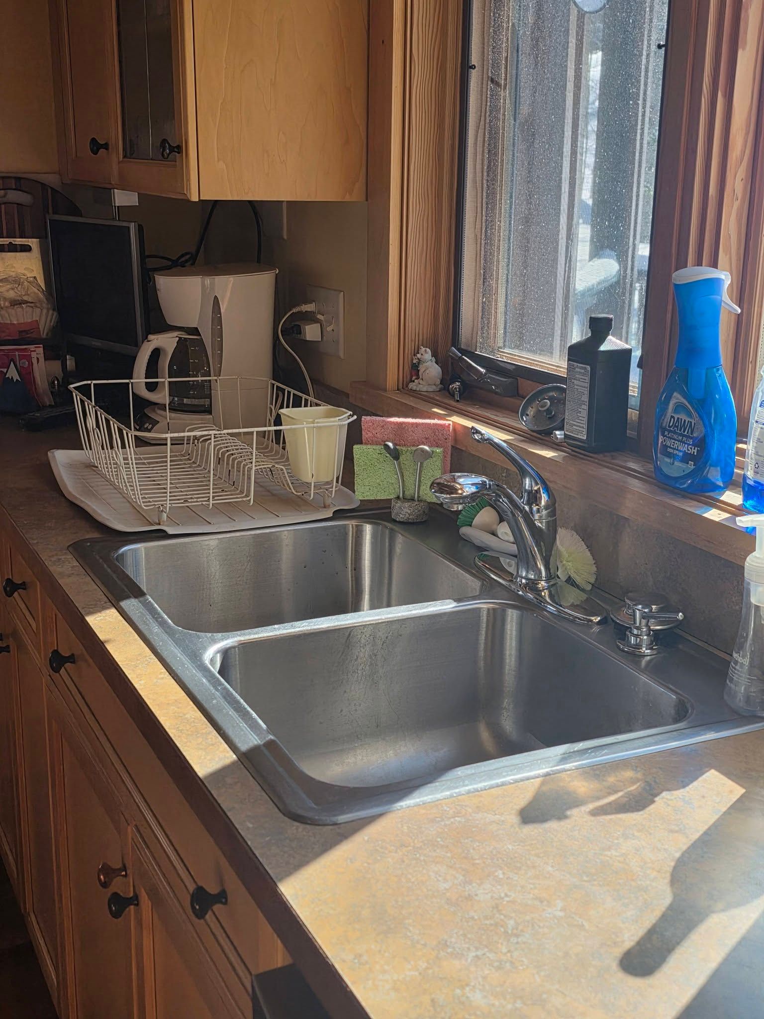 Sunlit kitchen sink with double basins, wooden cabinets, and dishes by a window.