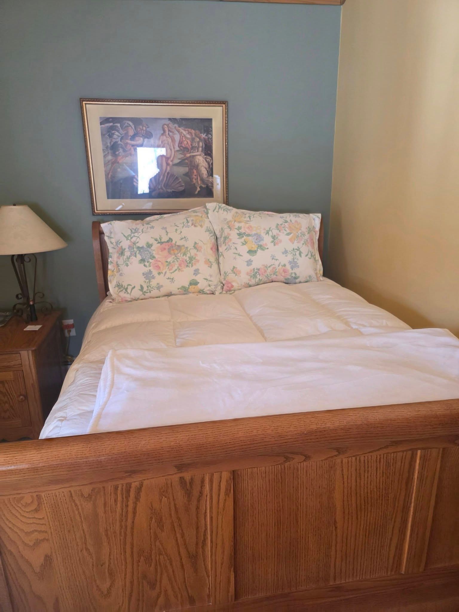 Wooden bed with white bedding and floral pillows in a small bedroom with a lamp and framed artwork.