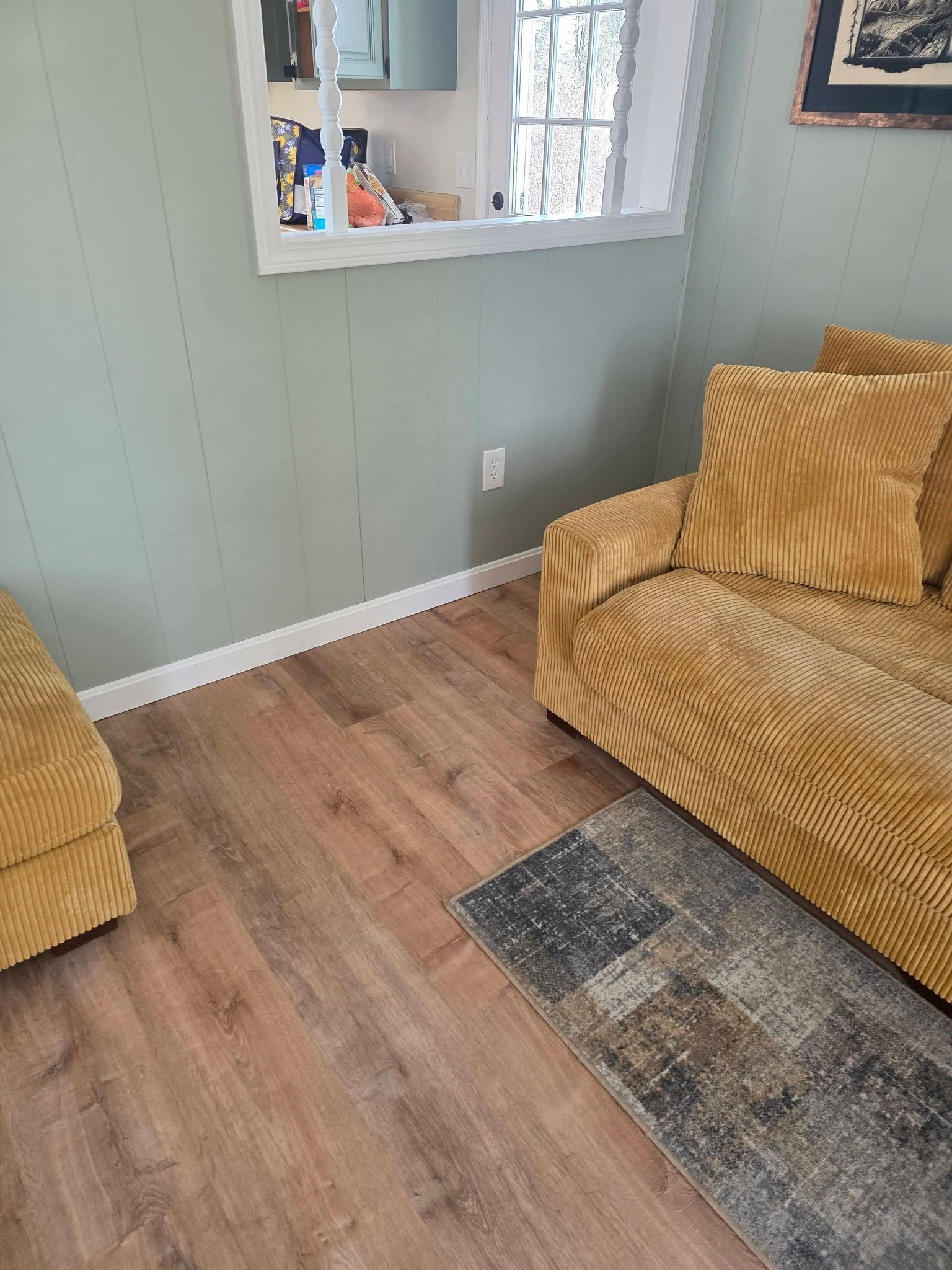 Small living room with sage walls, wood floor, tan couch and chair, window mirror, and a dark rug.