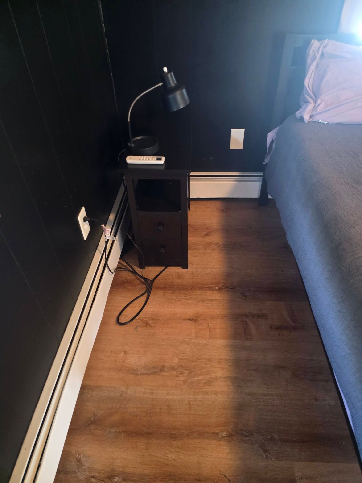 Small bedroom corner with a black side table, lamp, wall outlet, and bed beside a wood floor.
